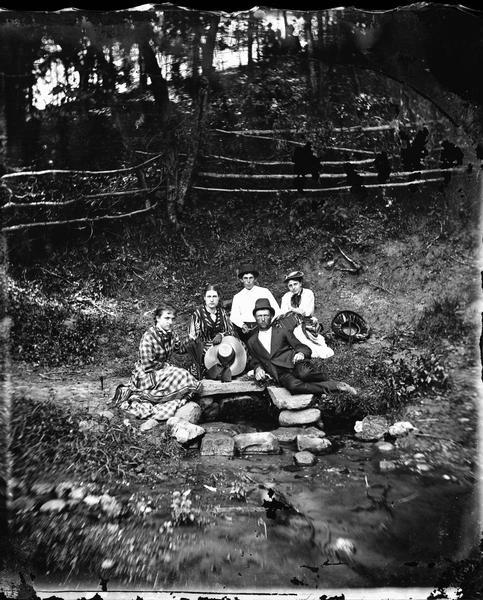 Group of people sitting beside spring, perhaps at Winnequah or Picnic Point in Madison.  All of them are either holding or are wearing hats.