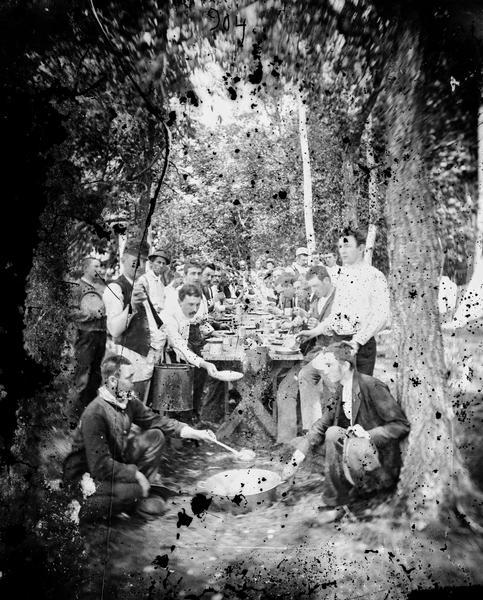 Chicago Traveling Club, trip #8, to Twin Lakes, Wisconsin. The campers are seated at a long table preparing to eat an outdoor meal.