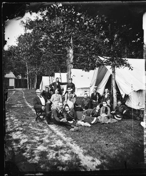 A group of interestingly dressed men and women posed before a line of tents. The men are wearing uniforms, possibly those of a band.  Fishing poles and chairs are behind them.