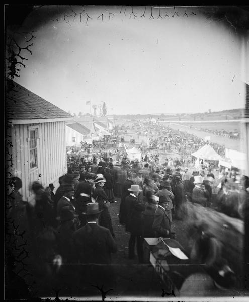 Crowd at the Camp Randall fairgrounds watching a horse race, part of that year's Wisconsin State Fair. Carriages and people are near buildings on the left, with the track, windmills and the U.S. flag in the background.