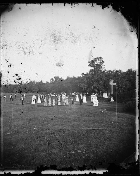 Large group at Syttende Mai (May Celebrations), possibly at Winnequah. Norwegian ladies are playing croquet and there is a Norwegian Flag on the side. Women hold open parasols and there are men on the side watching.