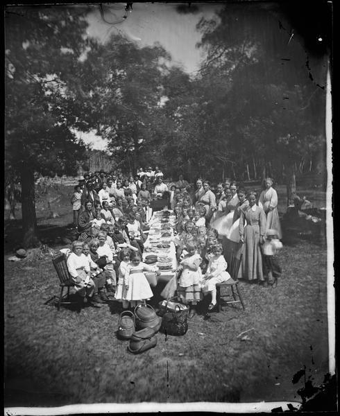 A large group of children are seated at a long table loaded with desserts. The boys sit on the left and girls on the right. Many are clutching what appear to be diplomas. Women stand nearby, and a mixed group of women and men are in the background. All are wearing fancy clothes. Straw baskets sit on the ground in front of the table.