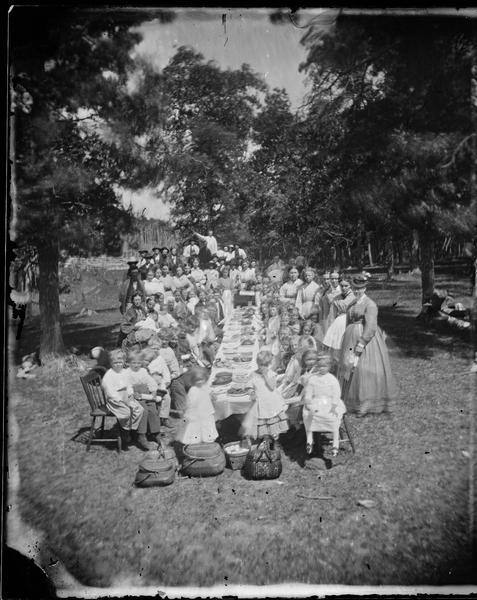 A large group of children are seated at a long table loaded with desserts. The boys sit on the left and girls on the right. Many are clutching what appear to be diplomas. Women stand nearby and a mixed group of women and men in the background. All are wearing fancy clothes. Straw baskets sit on the ground in front of the table.