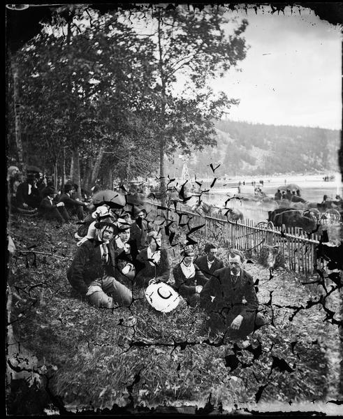 Well-dressed young couples sit in the foreground. In the background is a picket fence running along a busy road with horse and buggy traffic. Further on is Devil's Lake and a windmill. The occasion is the Grand Regatta held June 21-22, 1877.