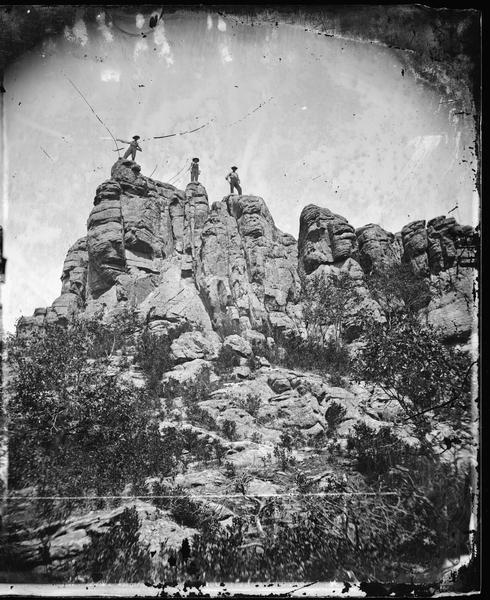 Three men standing on a rock formation possibly in the Baraboo Bluffs.