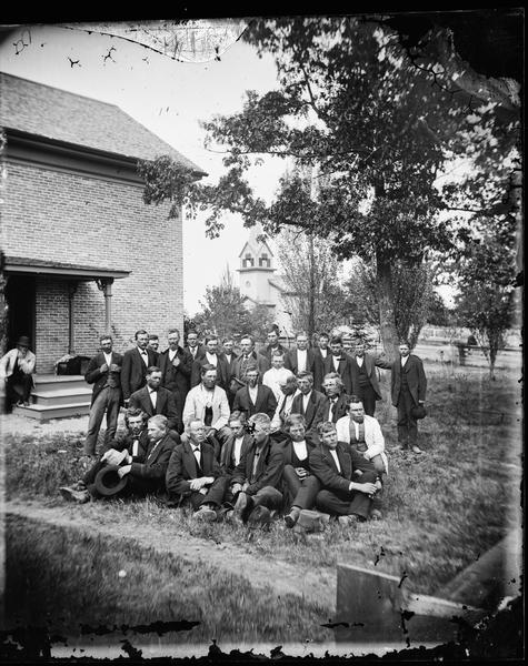 Group portrait of delegates to the meeting of the Eastern District Norwegian Synod, held at the Immanuel Norwegian Evangelical Lutheran Church, May 31-June 6, 1877. Immanuel Evangelical Lutheran Church can be seen in the distance. The view is from the front of the parsonage.