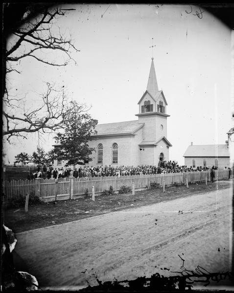 View across road of the Eastern District Norwegian Synod, May 31-June 6, 1877, at the Immanuel Norwegian Evangelical Lutheran Church (2 1/2 miles west of Larsen). The church was built in 1872. Pastor Even Johnson Homme is in the front row, left, of the group standing on the steps.