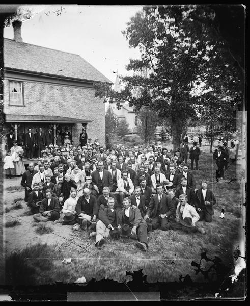 Group portrait in the yard of the Immanuel Norwegian Evangelical Lutheran Church parsonage. Pastor Even Johnson Homme and his wife Ingeborg and their children are in the front, center and left. Even Homme is on one knee, seated center front. The group is possibly the Winchester congregation. Photographed during the meeting of the Eastern District Norwegian Synod.