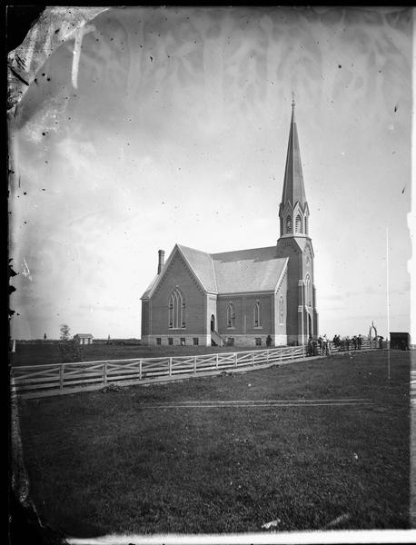 View down road towards the exterior of a large brick church with a stone foundation, belfry and steeple. A group of people stand in front of the white board fence in front of the church.