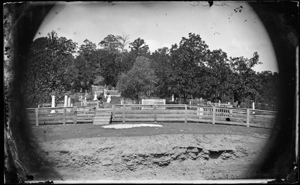 Two woman sitting in Oak Hill Cemetery. Board fencing, with a set of steps near the gate, surrounds the graveyard. Picket fences set off some of the headstones. The area in the foreground appears to be excavated below the grass.