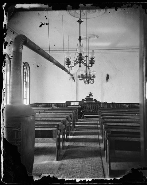 View of the primitive Albion Prairie Methodist church looking down the aisle between the pews towards the pulpit and a small organ. A prominent stove pipe runs the length of the church. A hemp runner is on the aisle and attractive oil lamps hang from the ceiling.