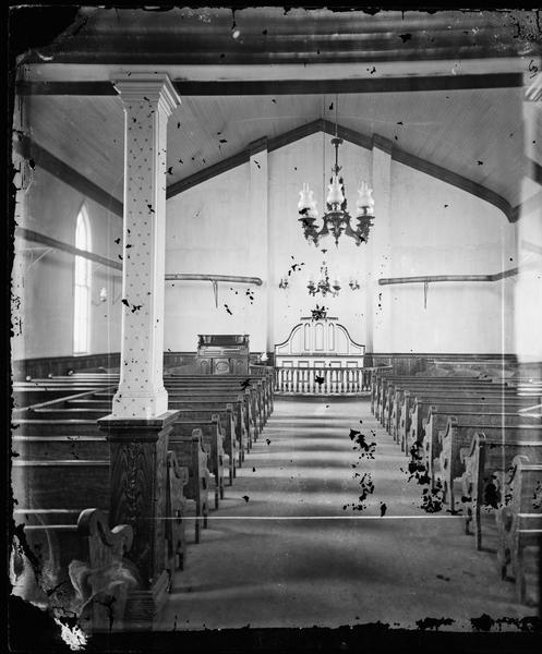 View of a church interior looking up the aisle between the pews towards the altar and a small organ. Twin stove pipes run along the side walls.