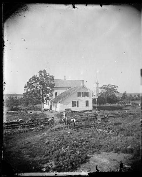 Men sit on farm machinery at the side of the parsonage of the St. Paul Liberty Church.  The church is visible in the distance. The town of Deerfield is found in the Liberty Prairie settled by Norweigan immigrants.