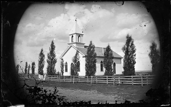 A frame church is enclosed in board fencing. Lombardy poplars line the fence.