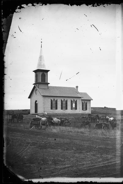 View of the Arlington Prairie Lutheran Church, a brick building with handsome shuttered Gothic windows.  The congregation's horse-drawn vehicles are assembled outside.