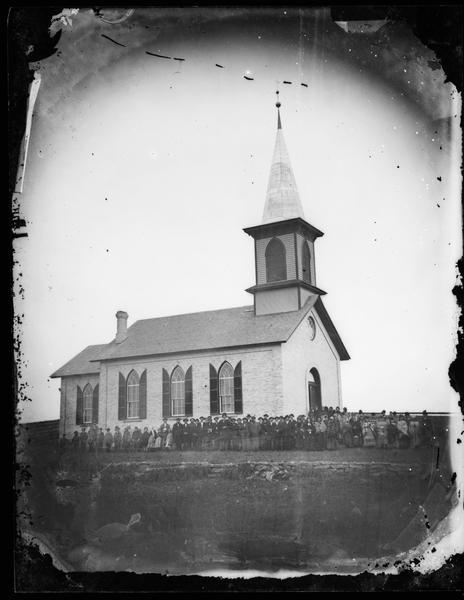 The congregation of the Arlington Prairie Lutheran Church assembled at the side of the church and dressed for the cold.  The church is a brick structure with shuttered windows.  A circular stained glass window is above the entrance.