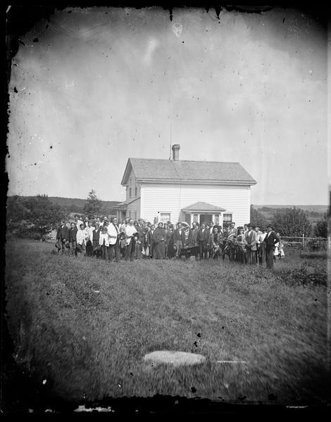 A funeral party is assembled around a casket in front of an upright-and-wing frame house.  Two older men on the left, one identified as Lars D. Reque, stand with Bibles while several women wearing Norwegian-style patterned shawls are standing close to the casket.  This house was insured by the Hekla Fire Insurance Company, which sold insurance to many Norwegian-American households in south central Wisconsin.