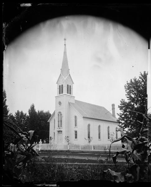 View from cornfield towards the Orfordville Lutheran Church, built in 1871-1872, surrounded by a white picket fence. The pastor at the time of the photograph was Johannes Muller Eggen (1841-1913).