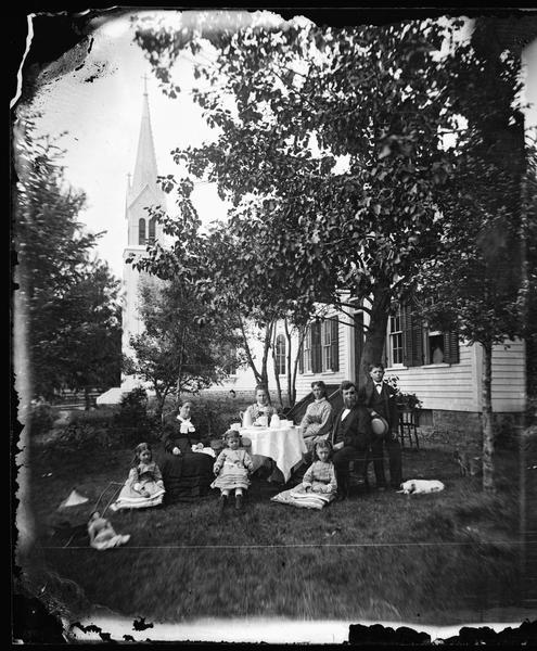 Pastor Claus Friman Magelssen (1830-1904), his second wife Marie Schlambusch Magelssen, and their family sit around a table set for coffee in the foreground.  The parsonage and the Luther Valley Church, built in 1872, are behind them. Visible are the minister's pipe, a baby carriage, a girl's doll and a dog.