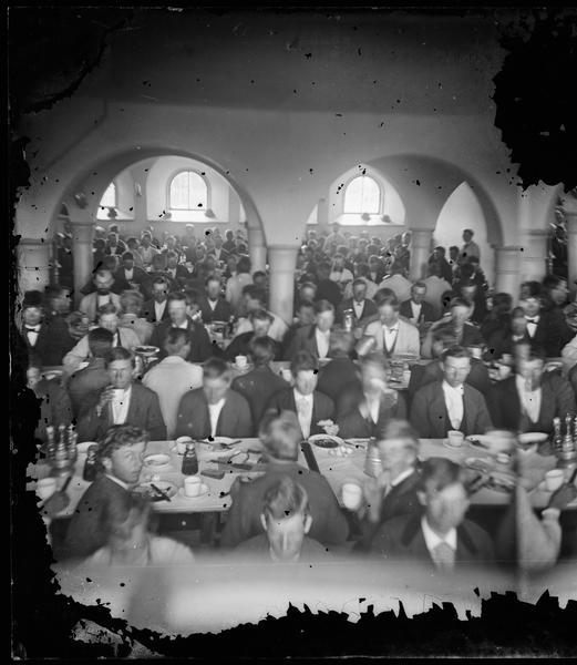 Elevated view of the Luther College dining hall interior. A large group of male students are seated in the hall which has arched windows and walls.