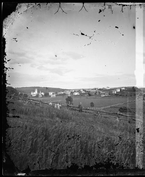 View of New Glarus from a neighboring hill. A man, wearing a hat and suit, and five children, girls and boys, are posing along the fence in the foreground. Fences surround the fields behind the group, and houses and church buildings are in the background.