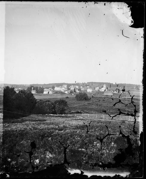 View, from a distance, of New Glarus. A hill, trees and fences are in the foreground.