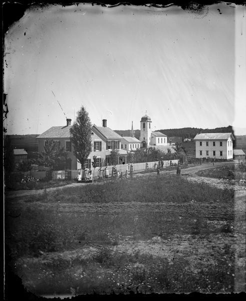 View of the town which was founded by Swiss settlers in 1845.  A group of women and children stands in front of a fence at 130 5th Avenue, the Gabriel Schindler house.  Newly planted trees line the fence.  In the background are two churches. The tallest church is the Swiss (Zwingli) Reformed Church, which was demolished in 1899.