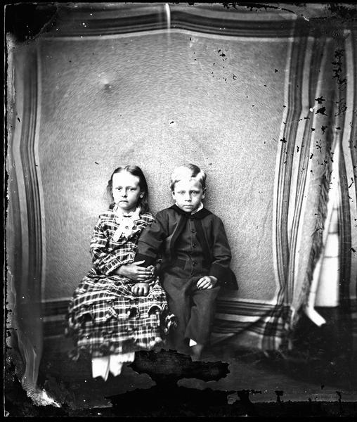 Seated indoor portrait of boy in checked suit and girl in plaid dress, perhaps taken at Dahl's DeForest studio.
