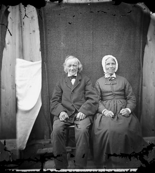 Portrait of older man and woman. They are sitting out doors side-by-side in front of a dark colored backdrop.