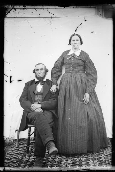 Portrait of Mr. and Mrs. Hans O. Hawkas of Bonnet Prairie Lutheran Church taken outdoors in front of a canvas backdrop. There is a carpet on the ground. Mr Hawkas is sitting in a chair, and Mrs. Hawkas stands on his left with her hand on his shoulder. Mrs. Hawkas is wearing a hoop under her skirts.