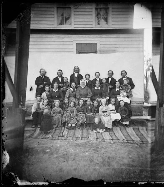 Group portrait of a family group of thirty men, women and children seated on steps against a cloth backdrop that has been attached to a frame house.