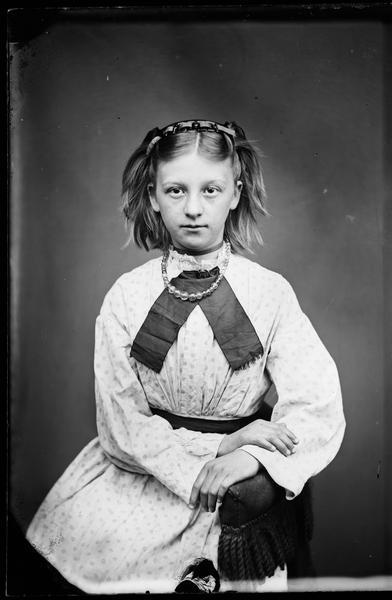 Seated studio portrait of a young girl. She is wearing glass or amber beads.