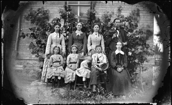 Outdoor family portrait with most of the girls dressed in the same fabric with vertical stripes. They are posed before a brick house.