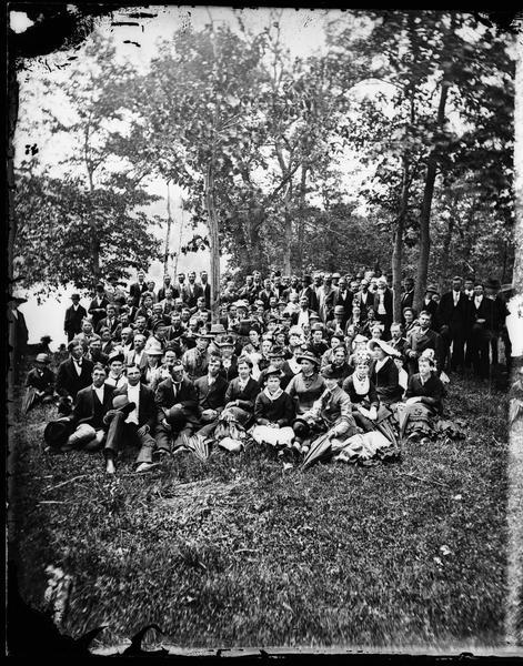 A large well-dressed group of men and women, with a scattering of children, posed outdoors near a lake. This group portrait may be at Maple Bluff on Lake Mendota near Madison.