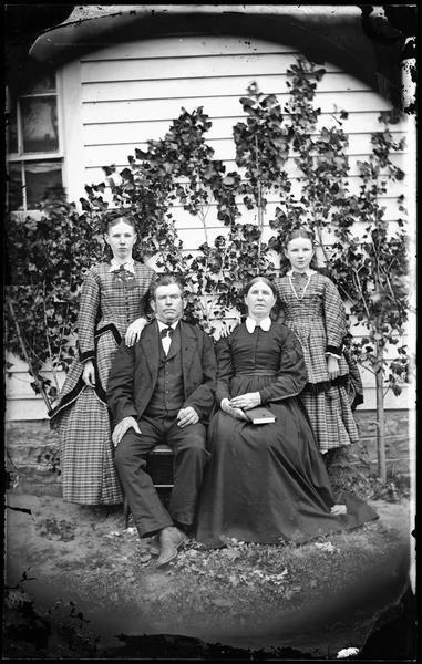 Group portrait of a husband, wife and two daughters, whose dresses are of different designs but made of the same plaid fabric. They are posed in front of a frame house.