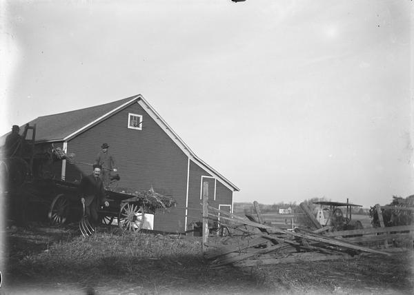 Three men with a wagon are on the left, with a barn, broken fence and farm implement on the right.
