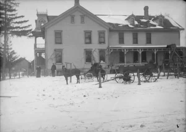 The Park Hotel, once lived in by Lewis Dahl and family. It was also the Isaac DeForest home. Men and women are standing outdoors. There is a wagon or carriage outside and snow on the top of a frame building. Two narrow windows on third floor of building, side porch and front porch with second story balcony tower type roof.