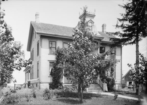 Isaac DeForest home. Pauline Dahl standing at the bottom of the front steps of large two-story frame house; the house has lattice work at bottom of porch, a railing at top, and a tower type structure on roof of porch. Carriage house in background on right. Converted to Park Hotel in 1890's by Ole S. Holum. Sold to Lewis Dahl in 1906(?).