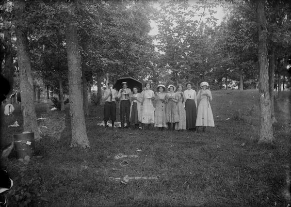 Outdoor portrait of nine people at Walter Dahl's class reunion. One person on the left is holding an umbrella. The group is standing together under trees holding plates and eating food; the women are wearing hats.