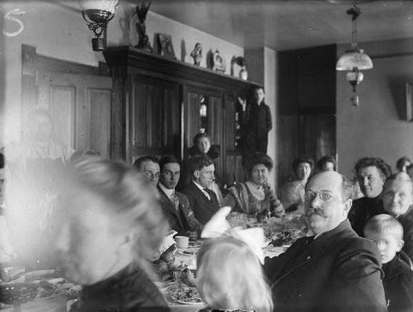 People around dining table that has food on it. A boy in the  background is standing on a chair.
