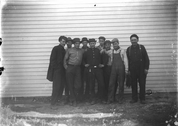 Eight men with hats, posed in front of a frame building. WED at bottom of negative.
