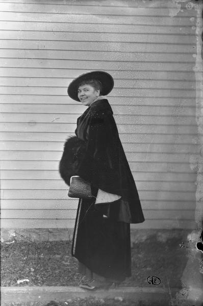 Woman outdoors in front of frame structure wearing a hat and cape, and holding a fur over one arm. GED initials at bottom.