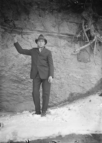 Walter Dahl standing outdoors wearing a suit and hat. He is standing in the snow, and behind him is a rock or dirt wall with tree roots sticking out of it. He is holding onto tree roots with one hand.