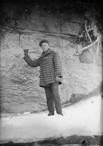 Mort Petit wearing a plaid coat and hat outdoors holding onto a tree root. Behind him is a rock or dirt wall with tree roots sticking out of it. Snow is on the ground. Mort Petit was Lewis Dahl's hired man.