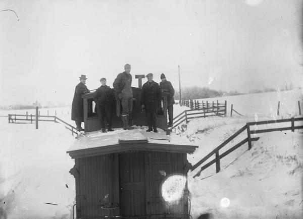 Elevated view of five men standing on top of the caboose. Heavy snow is on the ground, and fences are on the right and left.