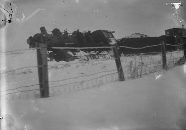View through fence looking towards the train wreck of "Soo Line". The engine is leaning off the track. Snow is on the ground.