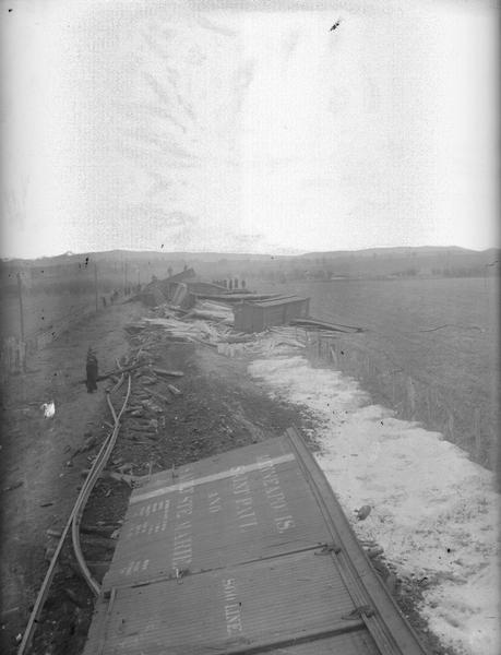 Elevated view of train wreck of Soo Line. There is a box car off the train tracks, with other wreckage in background. There is some snow along the right side.