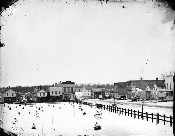 View of main street in Lake Mills. On the left of the image are the Fargo and Ostrander store, Farmer's Cash Store and Drugs, and A.J. Foster Harness Shop. Another harness shop and drug and hardware store are on the right.