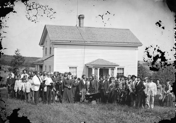 Group portrait of a large funeral party assembled around a casket in front of an upright-and-wing frame house. Two older men on the left, one identified as Lars D. Reque, stand with bibles while several women wearing Norwegian-style patterned shawls are standing close to the casket. This is another house insured by the Hekla Fire Insurance Co., which sold to many Norwegian-American households in south central Wisconsin.