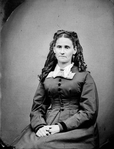 Seated portrait of a woman with curled hair, lace trim collar with pin and earrings.